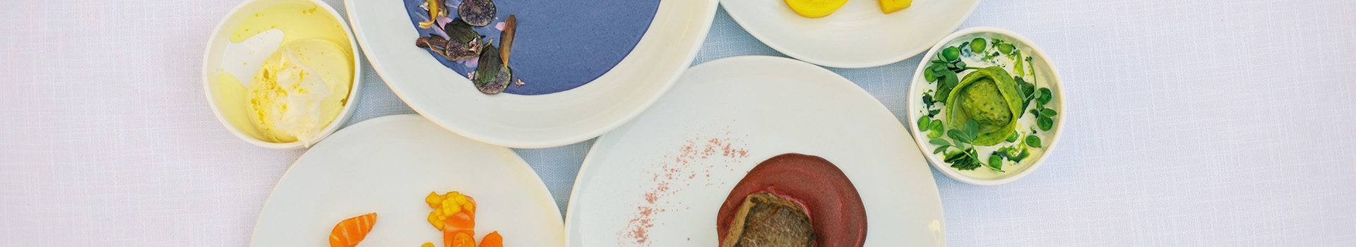 Overhead of white plates on a textured white background. From left to right, in the left top is a small bowl of ice cream, next to it large bowl of purple potato soup cut off, next to it a plate of yellow heirloom tomatoes, and a small bowl of a green tortellini. At the bottom left, is orange salmon and next to it is a red bison tenderloin with cherries three ways