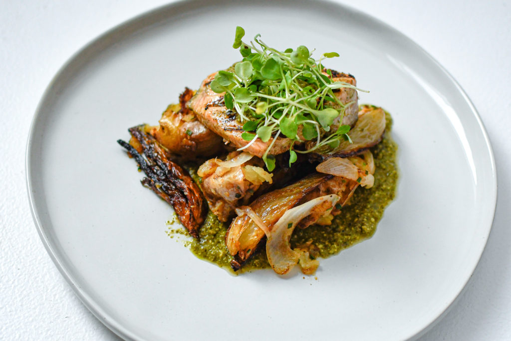 A piece of grilled salmon on top of roasted veggies with green sauce underneath. On top of the salmon is some microgreens. Everything sits of a white plate on a white textured background.