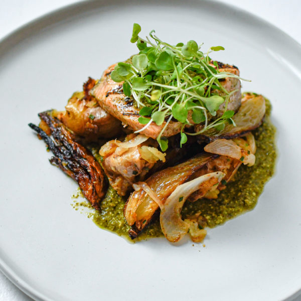 A piece of grilled salmon on top of roasted veggies with green sauce underneath. On top of the salmon is some microgreens. Everything sits of a white plate on a white textured background.