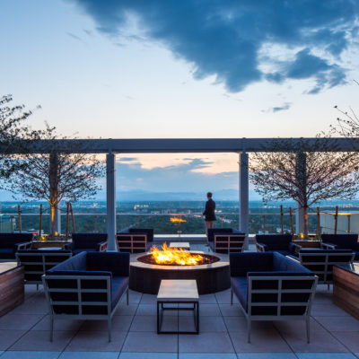 On the rooftop of Terrance at Colorado with a fire pit lit surrounded by outdoor lounge furniture. A man is looking off the balcony down at the city of Denver with mountains in the background