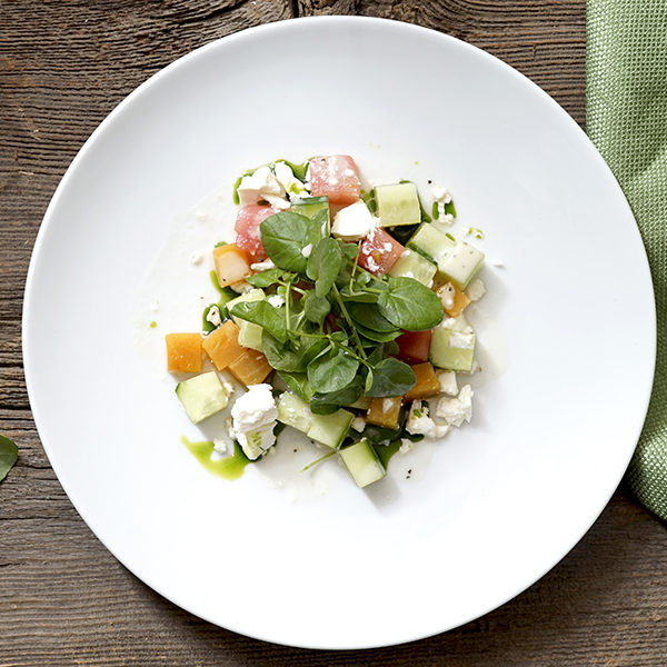 Overhead of the triple cube salad with cucumber, watermelon, feta, cantalope with a bit of greens on top on a white circle plate on a wood background