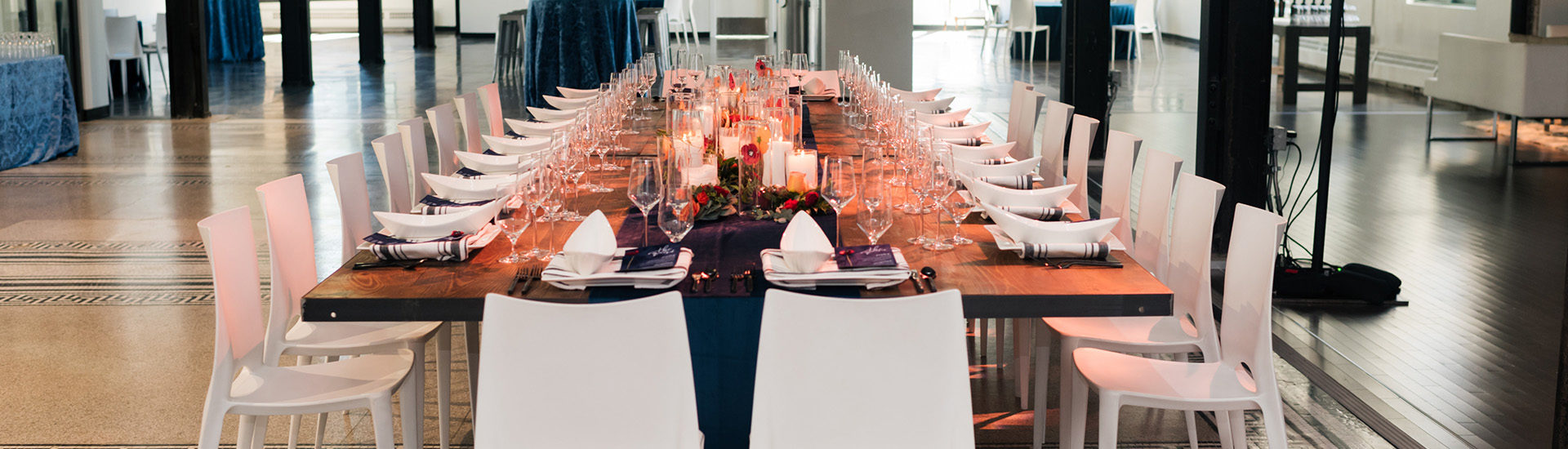 Close up of a long wooden table with modern white plastic chairs. At seating are a white glass, champagne class, and water glass, a rectangle plate with a blue and white striped napkin, oval white bowl, and navy menu. In the middle is a solid navy runner with paper flowers, and thick white candles are different heights.