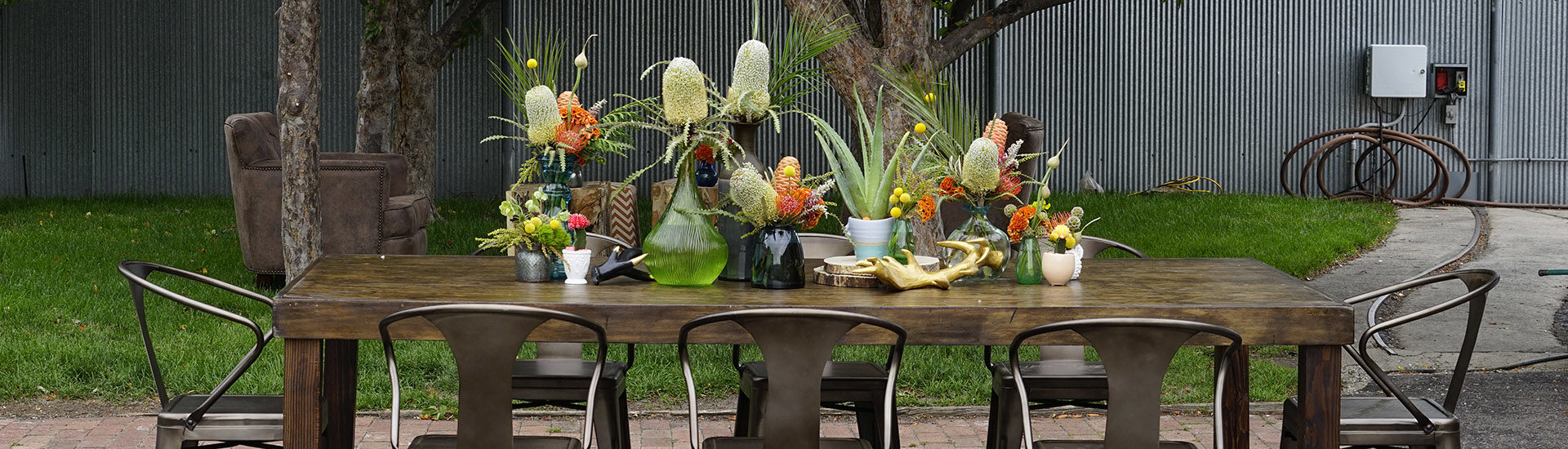 Long wooden table surrounded by metal chairs with a lot of florals in different colored vases at different heights