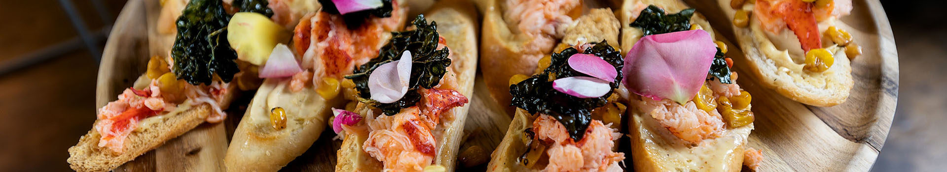 Slices of toast topped with lobster, topped with a crispy kale chip and flower petals. They sit on a wooden plate.
