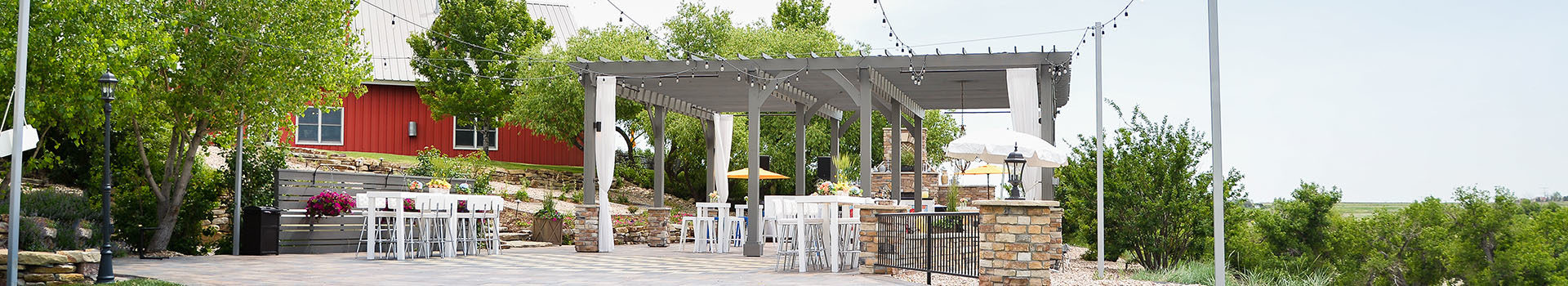 A grey pergola on a stone patio, white table tops with white barstool chairs and a red barn in the background with lots of trees and greenery.