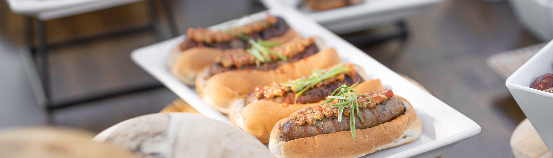 Close up of 4 brats in puns on a white rectangle platter topped with mustard a drizzle of ketchup and green onion