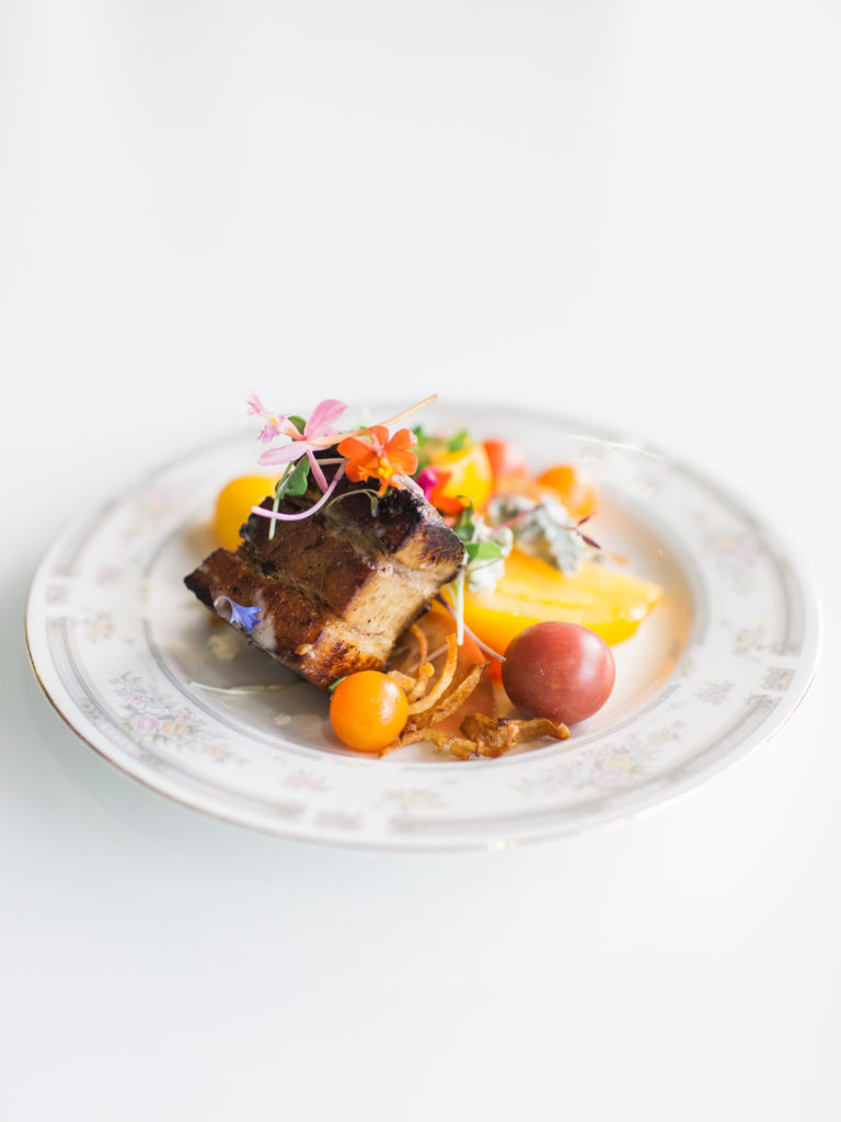 Close up of a slice of pork belly with cherry tomatoes and micro greens and edible flowers as garnish on an antique white grey plate