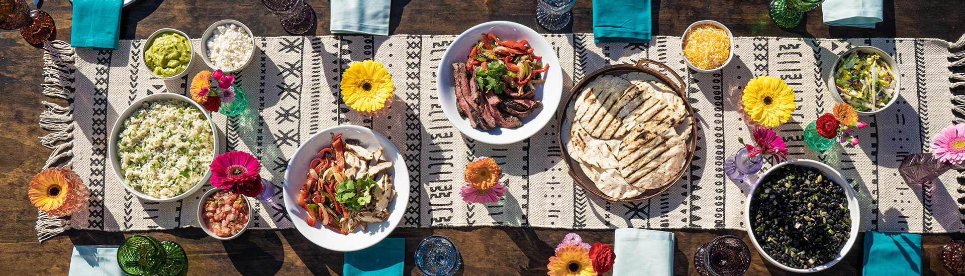 Overhead of different dishes for a fajita fiesta in white bowls. Underneath is a patterned black and light tan runner with fringe on the bottom. The table is decorated with colorful chalice classes, vibrant flowers in clear glasses and different shades of blue napkins.