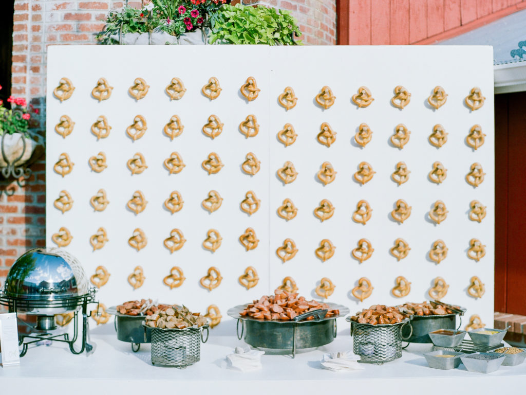 Twisted pretzels on a white wall with wooden pegs. Underneath it on a white table with different types of sausages sliced up on a silver display trays with 5 different sauces in small square bowls to the right.
