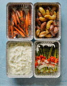4 aluminum rectangle boxes on top of a textured blue background. In the upper left box are roasted carrots, in the upper right are fingerling potatoes roasted, in the bottom left is a box of mashed potatoes and to the bottom right are roasted or grilled asparagus topped with sundried tomatoes, parmesan cheese chips and pine nuts.