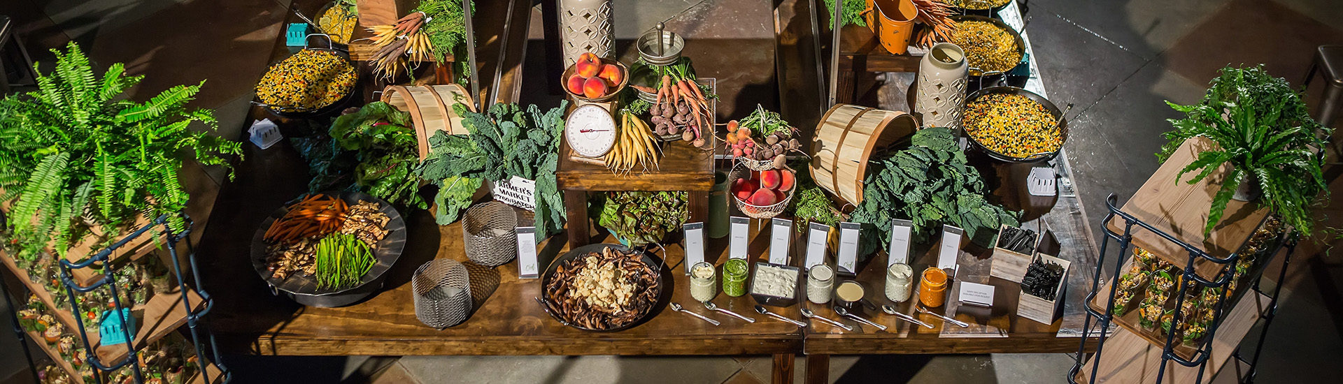 Overhead of wooden tables in a square with whole vegetables decorating the tables and sauces in mason jars, cooked vegetables and different types of corn in platters