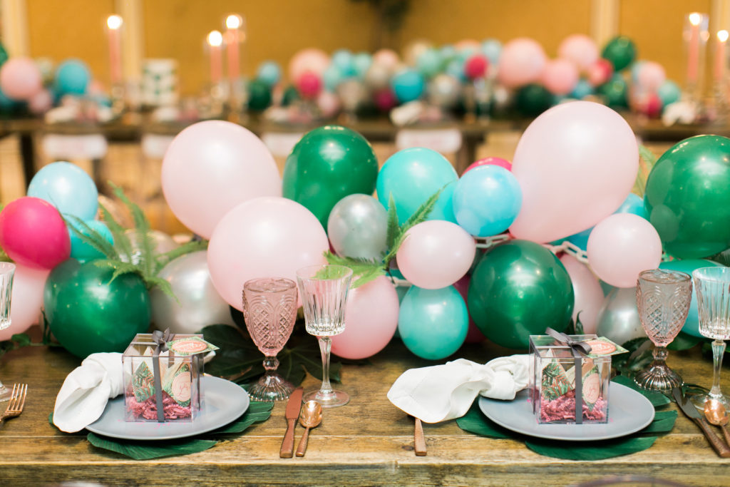 A rustic wooden table with a large long balloon garland with light pink, silver, emerald and turquoise balloons. On the table are the fortune cookie menu in a clear box sitting on a grey plate with a palm leaf placemat.