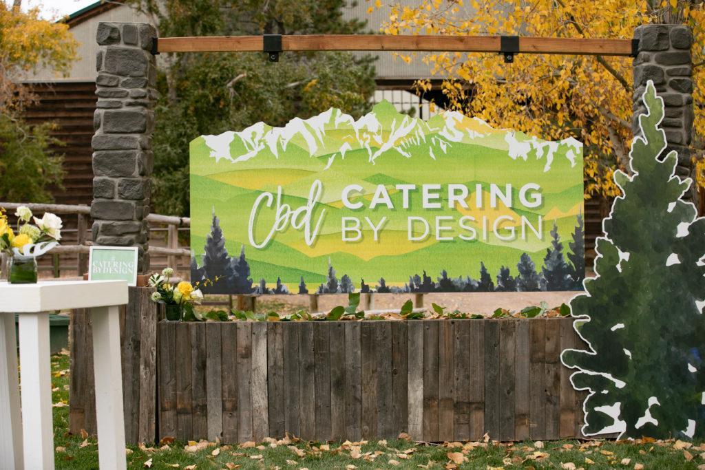 A large sign with a white Catering by Design Logo on a mountain sign with yellow and green
