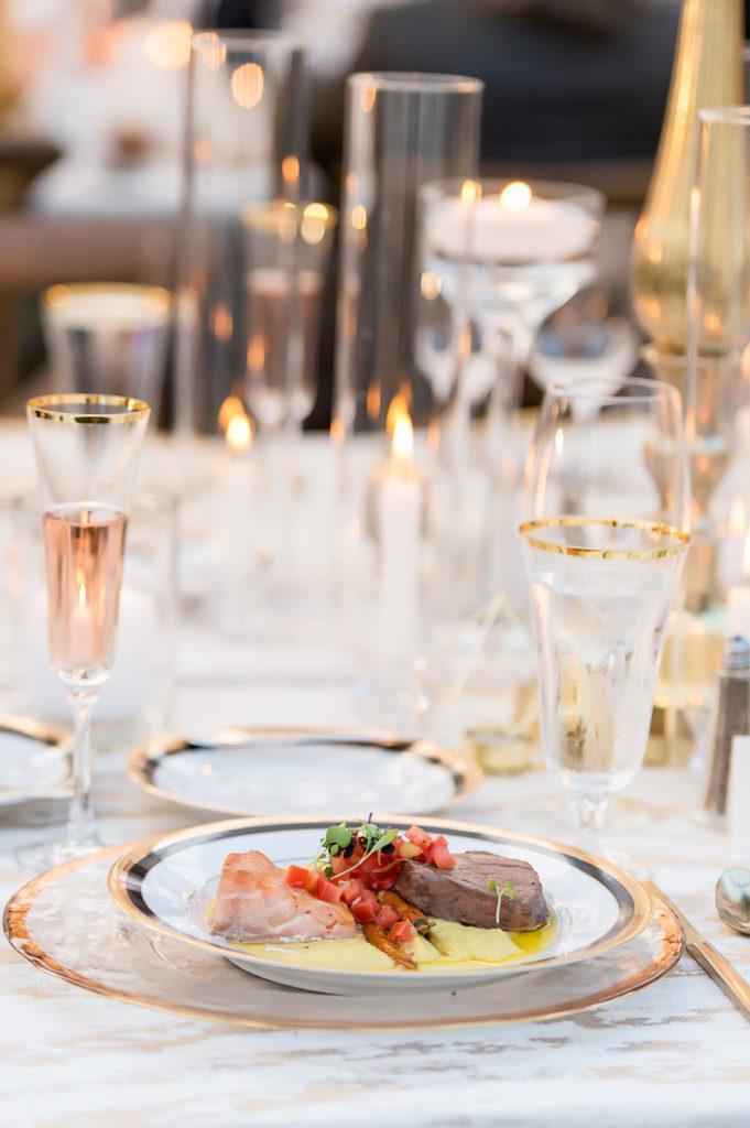 A place setting with rose gold accents around the edge on the plates and glassware. On the small plate are salmon and beef with tomato relish and potato puree.