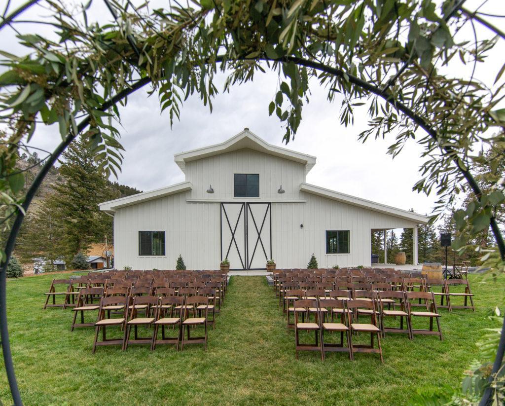 An outside ceremony with circle backdrop with greenery and brown folding chairs in front of a large white barn on green grass