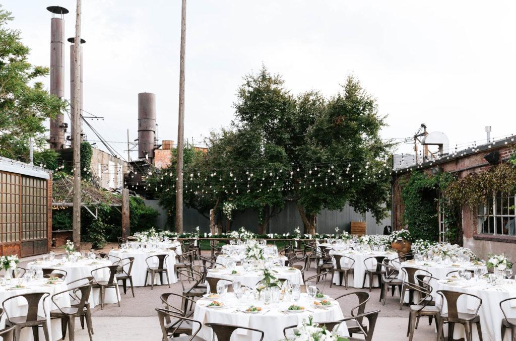 A bunch of circle tables outside in a courtyard with white table linens and metal chairs with a plated salad at each place seating