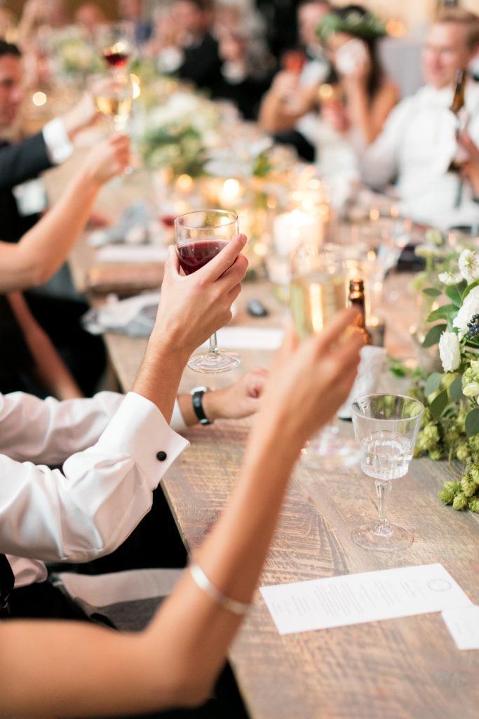 A wedding party sitting at a long wood tables holding either wine, champagne or beer to 'cheer'