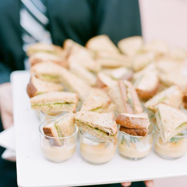 A white tray with small glass cups of couple with a mini sandwich held by a server wearing black and a stripped tie