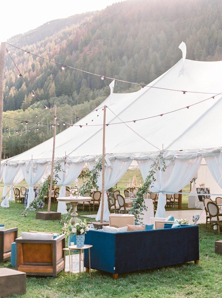 A giant white open tent outside, surrounded by the mountains. The tent inside is set up for a wedding reception and outside are luxurious couches and lounge furniture