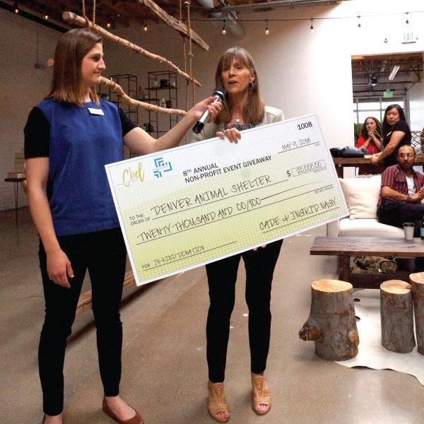 A women holding a giant check for $20,000 speaking into a handheld micrograph that another women in a blue polo and black pants is holding