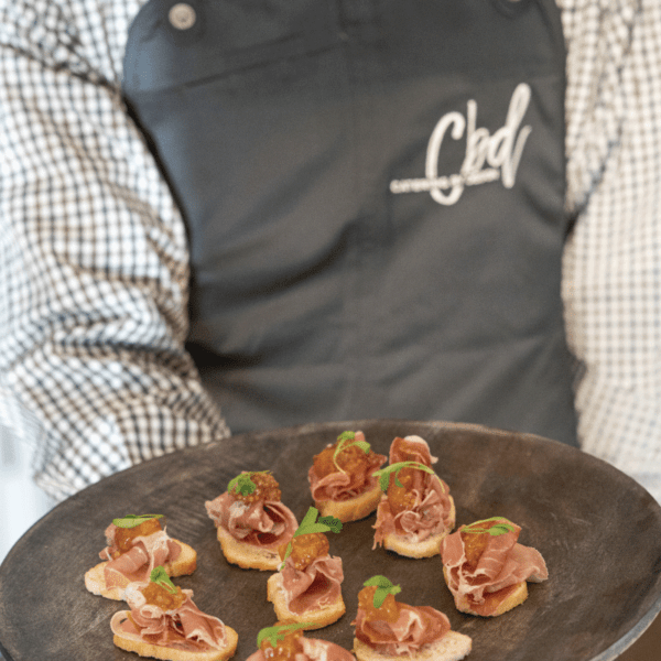 A server wearing a grey, brown and white button down with a slate colored apron with the Catering by Design logo in white is holding a circle tray with crostini with prosciutto