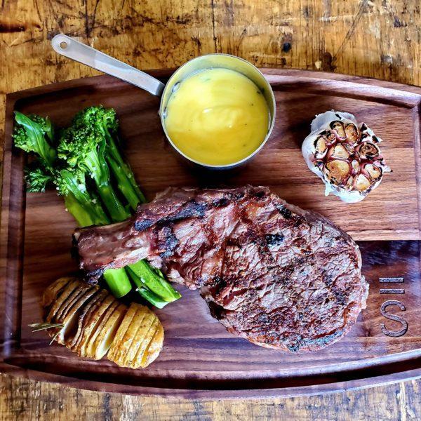 A tomahawk steak on a wooden board with a whole head of garlic, sauce, broccolini and hassel back potato surrounding the steak