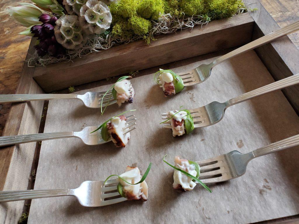 6 forks on a wooden tray. At the tip of each fork is a bit of octopus and greens.