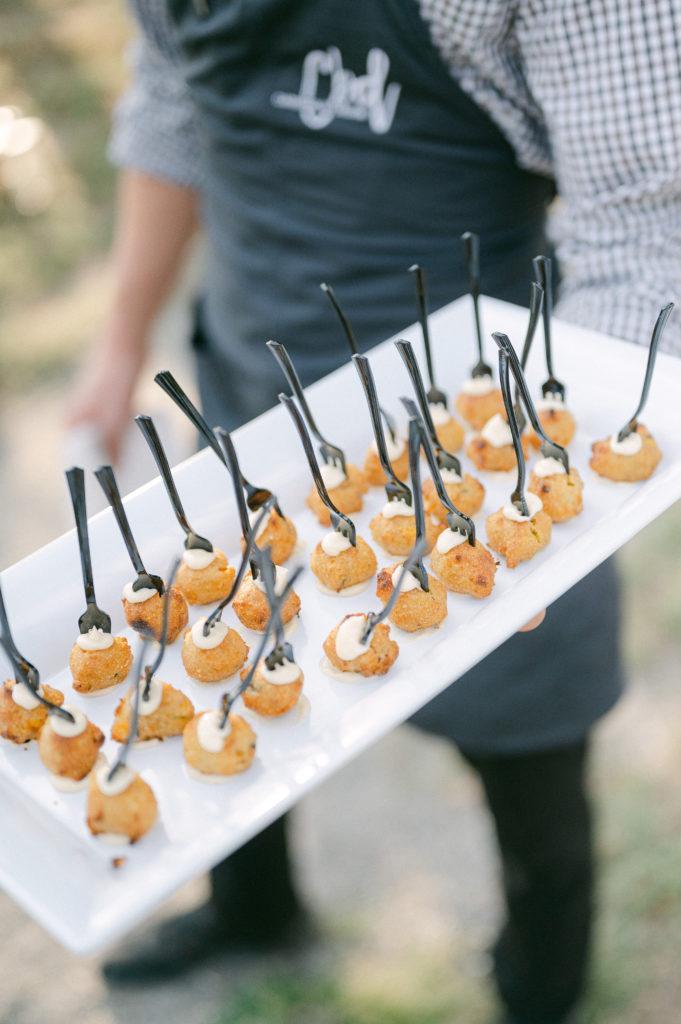 A small hors d'oeuvre with. a mini fork sticking out with a white sauce on top and bottom is held in the hand of a server.