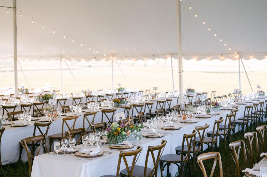 Multiple tables with white tablecloth and wood chairs are underneath a tent set up for an outdoor reception
