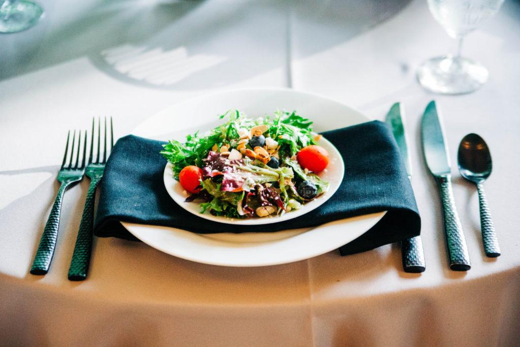 A hive salad on a small white plate sitting on a black napkin on a dinner plate. Dark chrome silverware is plated underneath a white tablecloth.