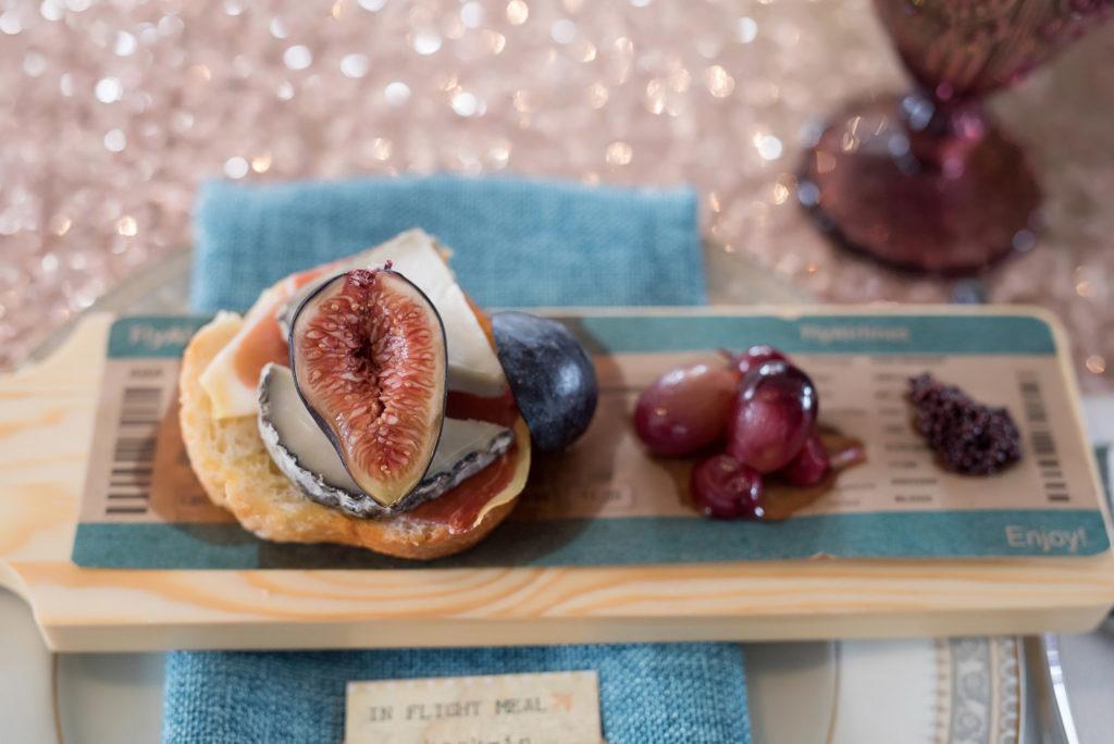 Fig crostini on a wooden paddle with an overlay of a plane ticket on a sparkly pink sheet