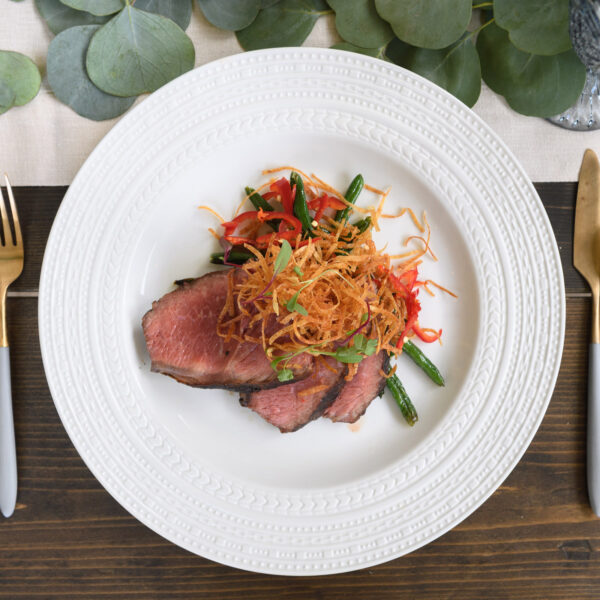 Overhead of a steak sliced on a white plate with green beans, peppers and frizzled sweet potato on top.