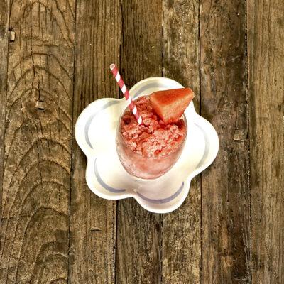 watermelon granita in a glass on a white flower shaped plate with a piece of watermelon and red and white striped straw