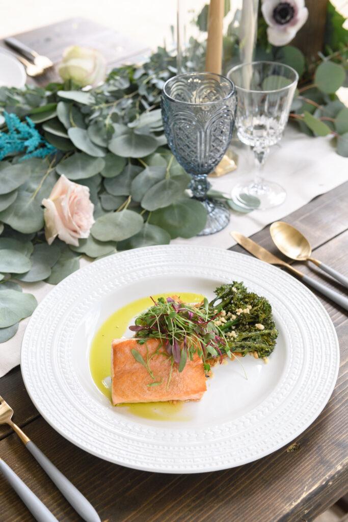 A white dinner plate with a piece of salmon with broccolini on a wooden table with greenery and flowers and grey and cold silverware