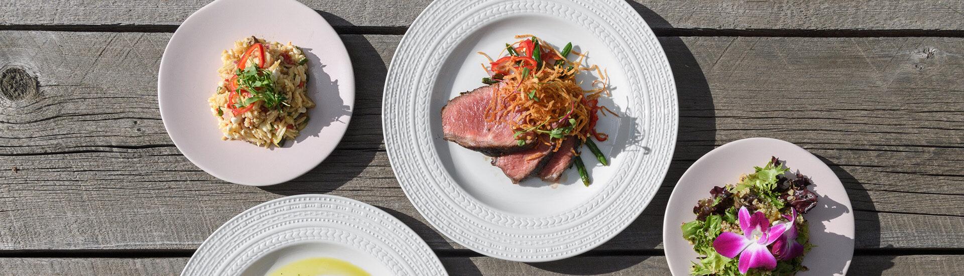 Overhead of multiple plates. A white dinner plate with sliced steak, a small pink plate with orzo salad, a small grey plate salad on a grey wood background