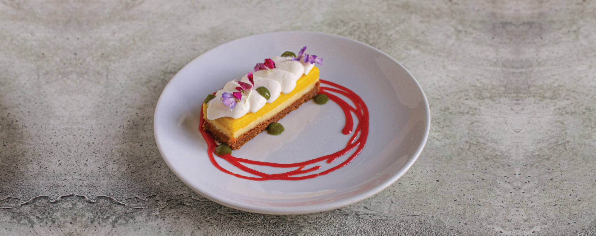 A slice of Lemon Basil Cheesecake on an off-white shiny dessert plate on a concrete background.