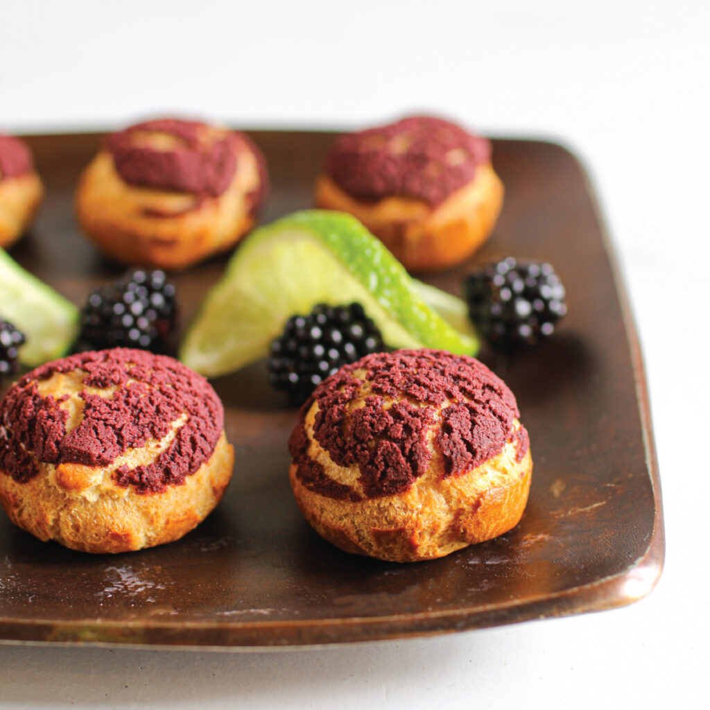 Creme puffs with a dark purple top with sliced limes and fresh blackberries on a copper plate.