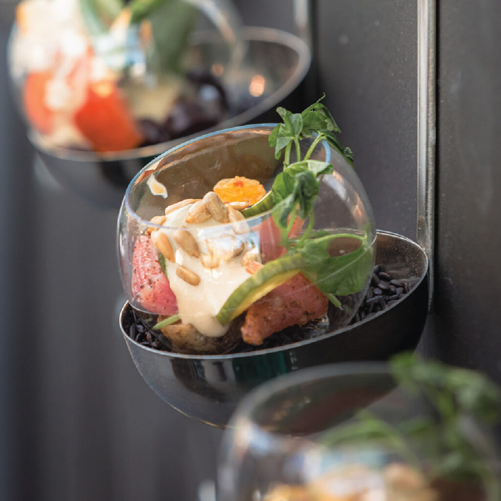 A mini salad in a plastic globe that sits on dry black rice in a ladle attached to a wall.