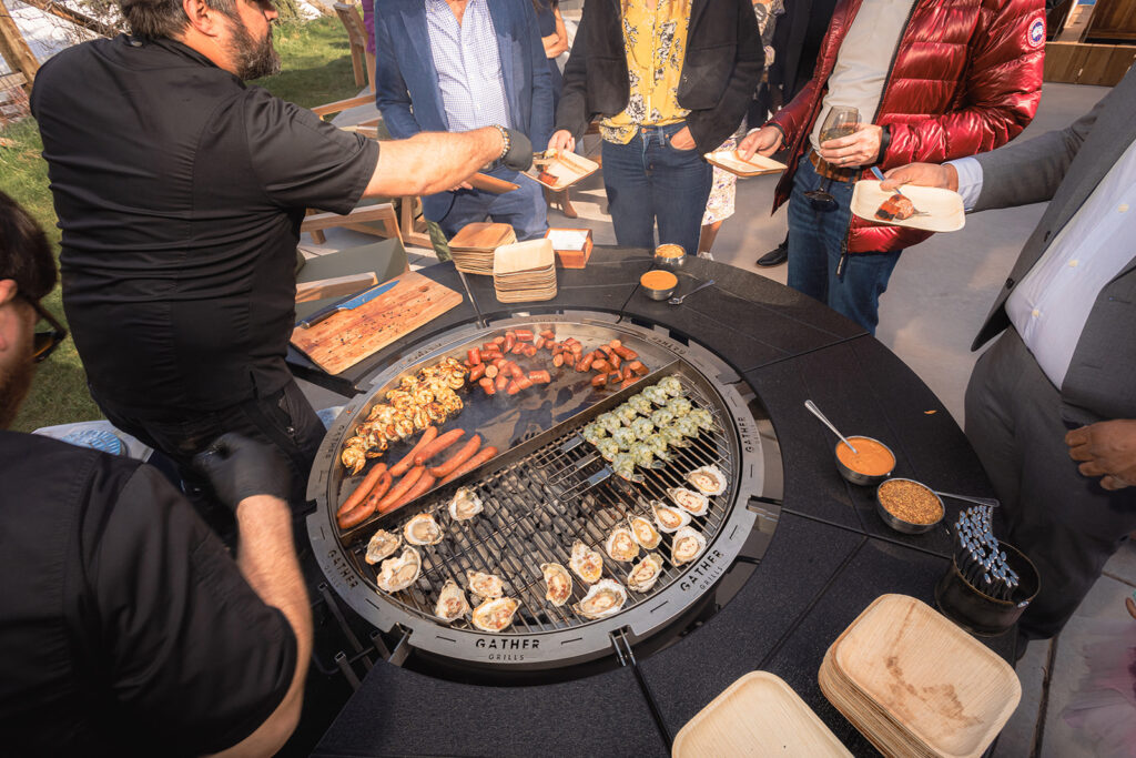 Various foods like sausage and shrimp being grilled on a gather grill with chefs and people surrounding the table