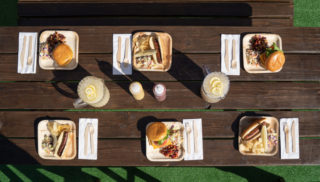 Square bamboo plates with either burgers or hot dogs with different picnic sides like cole slaw or potato chips on a pcinci table.
