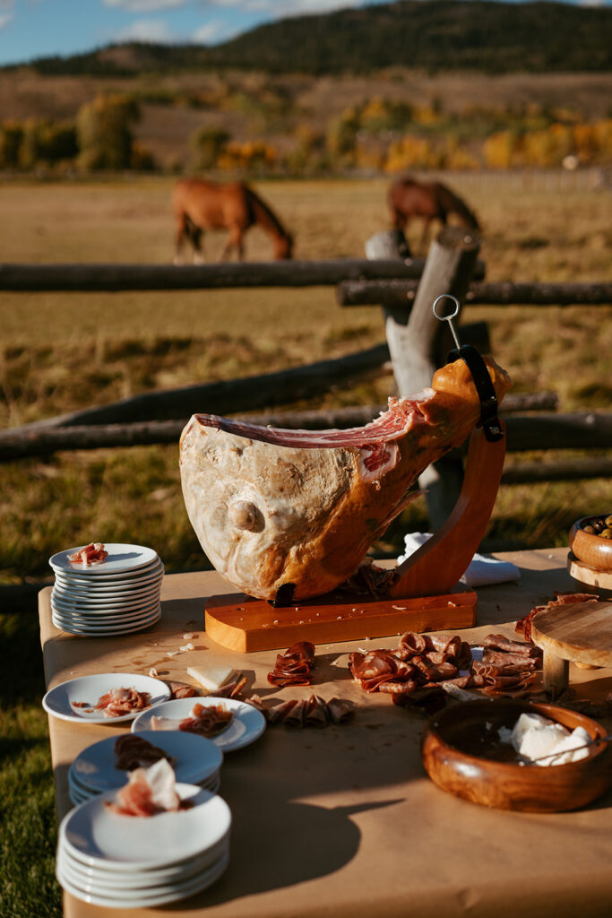 Prosciutto carving station with artisanal accompaniments overlooking a scenic Colorado ranch landscape.