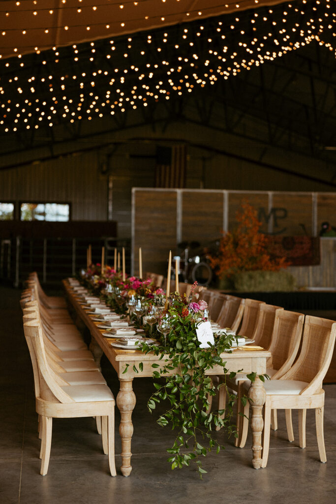 Long reception table with floral centerpieces beneath twinkling string lights inside a rustic barn.